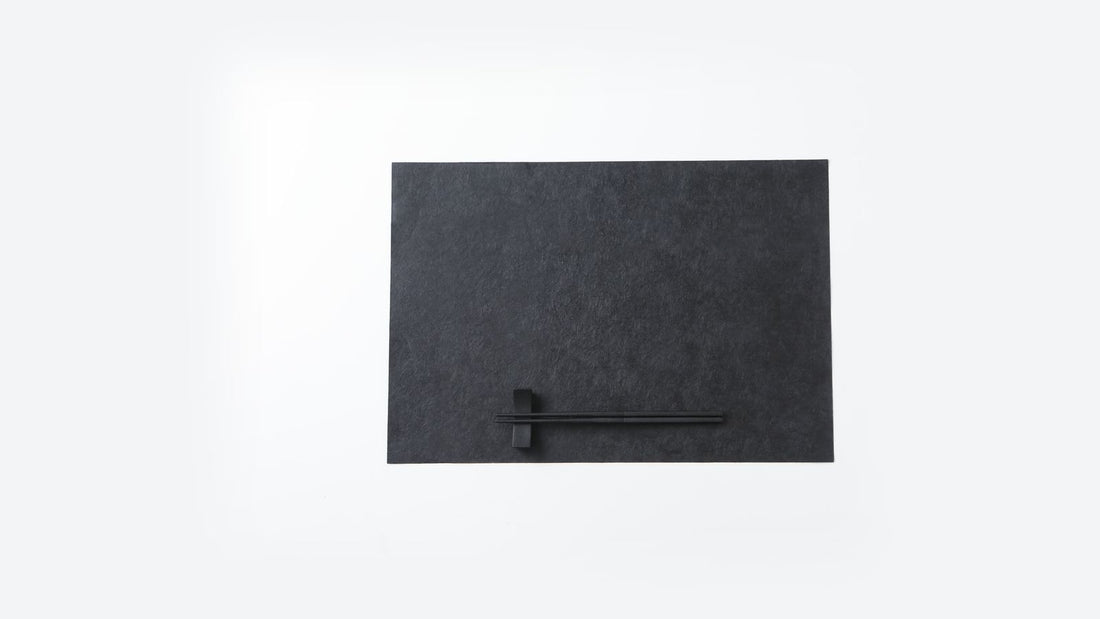 A dark urushi-finished washi placemat with black chopsticks laid on a chopstick rest