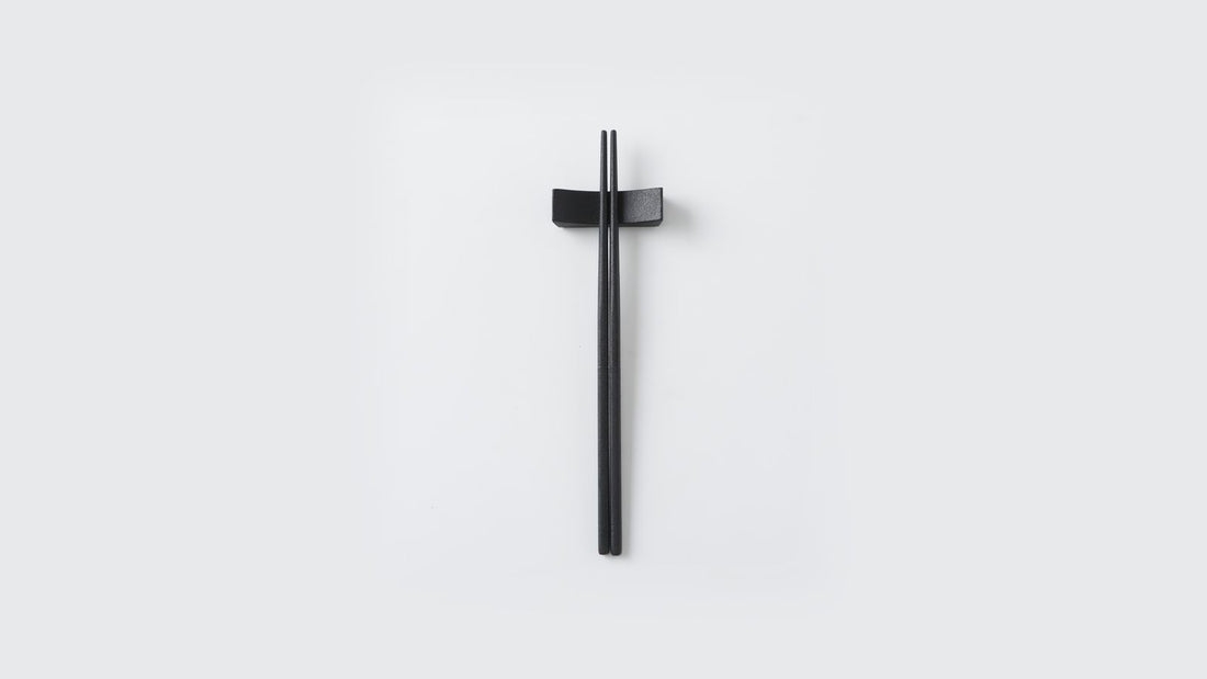 Black stainless steel chopsticks finished in urushi, resting on a matching chopstick rest on a light background