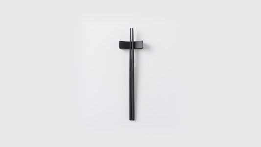 Black stainless steel chopsticks finished in urushi, resting on a matching chopstick rest on a light background
