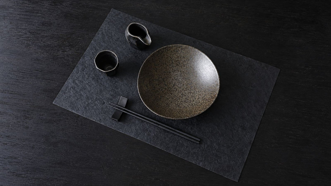 A Japanese place setting on a dark washi placemat: a ceramic bowl, two small cups, and black chopsticks on a rest