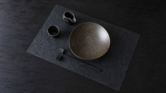 A Japanese place setting on a dark washi placemat: a ceramic bowl, two small cups, and black chopsticks on a rest