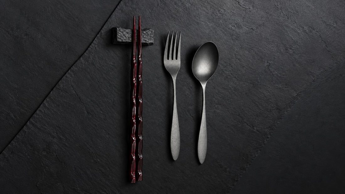 Wooden tame-nuri chopsticks on a stone rest beside an urushi-finished stainless steel fork and spoon on slate