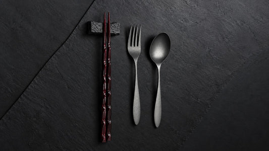 Wooden tame-nuri chopsticks on a stone rest beside an urushi-finished stainless steel fork and spoon on slate