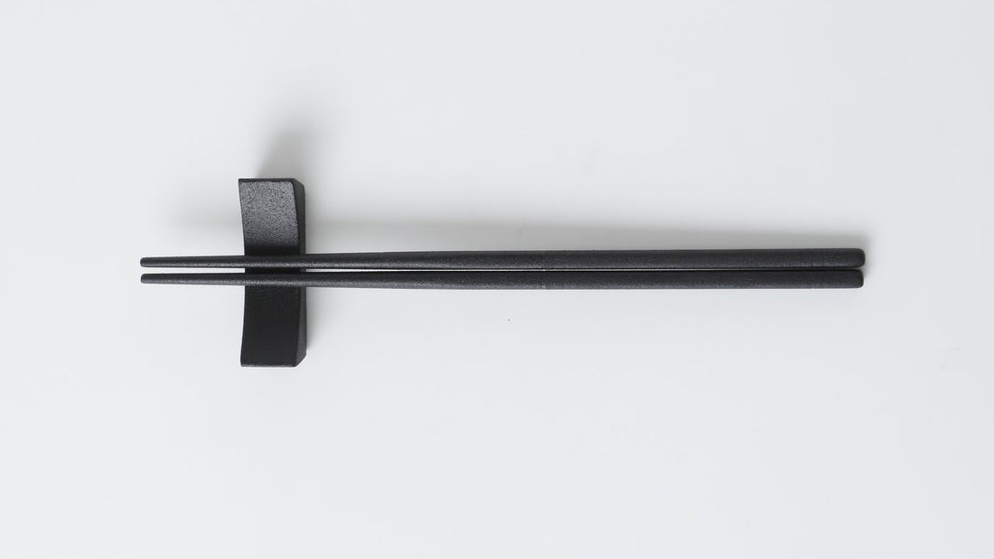 Black urushi-finished chopsticks lying horizontally on a chopstick rest with the tips pointing left, on a white background