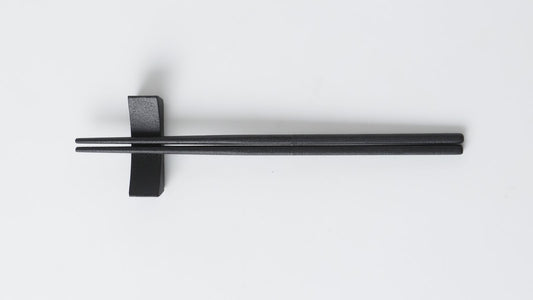 Black urushi-finished chopsticks lying horizontally on a chopstick rest with the tips pointing left, on a white background