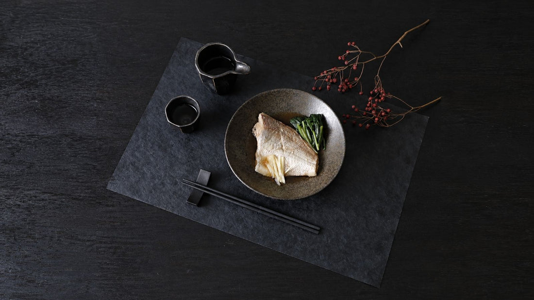 Japanese table setting on a black washi placemat: simmered fish in a stoneware bowl, sake cups, and black chopsticks on a rest in front with tips pointing left