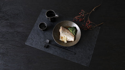 Japanese table setting on a black washi placemat: simmered fish in a stoneware bowl, sake cups, and black chopsticks on a rest in front with tips pointing left